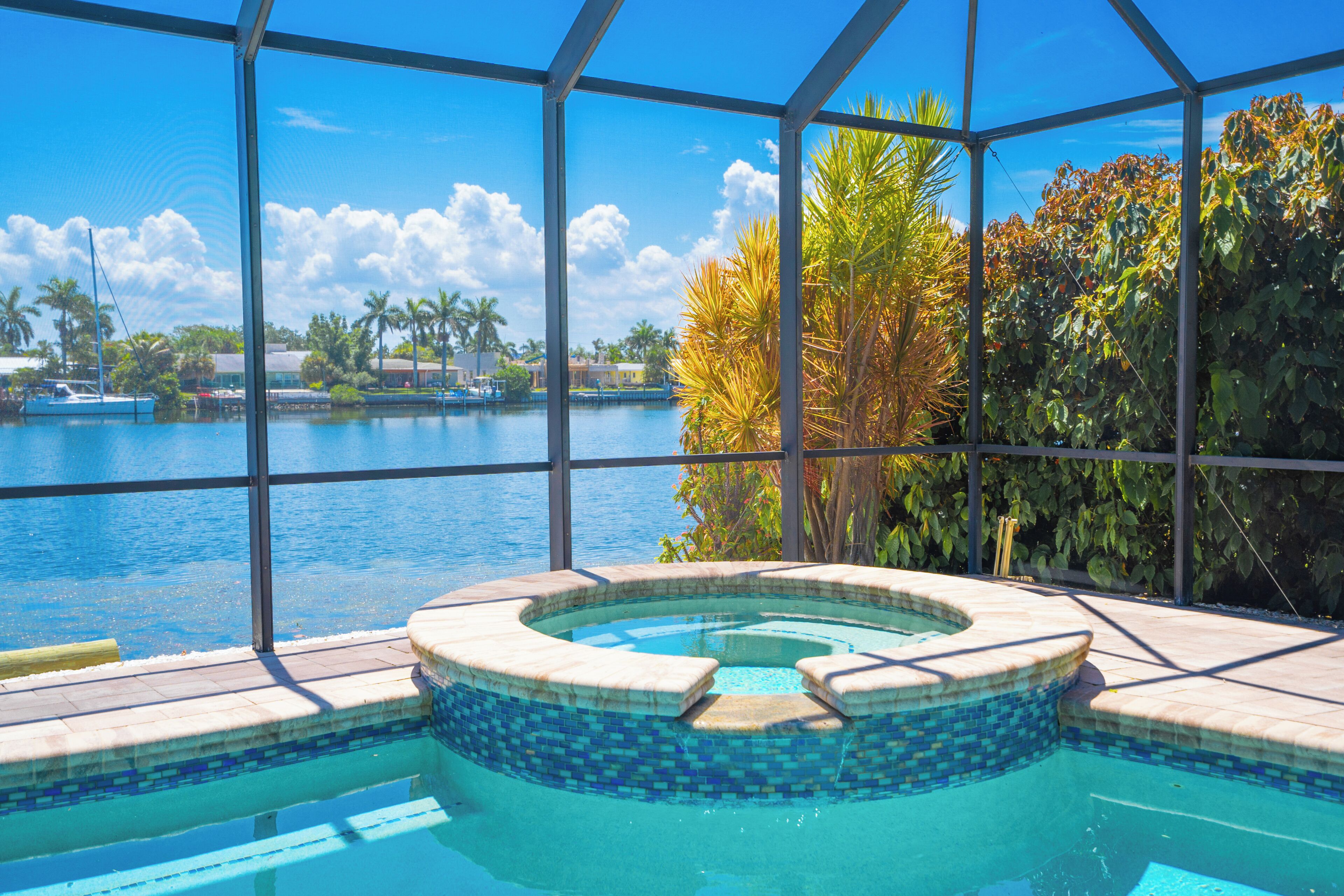 A pool with a hot tub, surrounded by a glass enclosure with a view of a lake and palm trees.