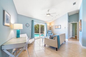 Un salon spacieux doté d’un ventilateur de plafond, un coin repas avec une table et des chaises, un canapé agrémenté d’un plaid, et une vue sur une piscine, visible à travers de grandes portes vitrées.