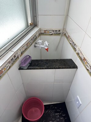 A bathroom with a window, a sink with a faucet, a purple cleaning brush, a red bucket, and a black mat with 'Coffee Time' written on it.