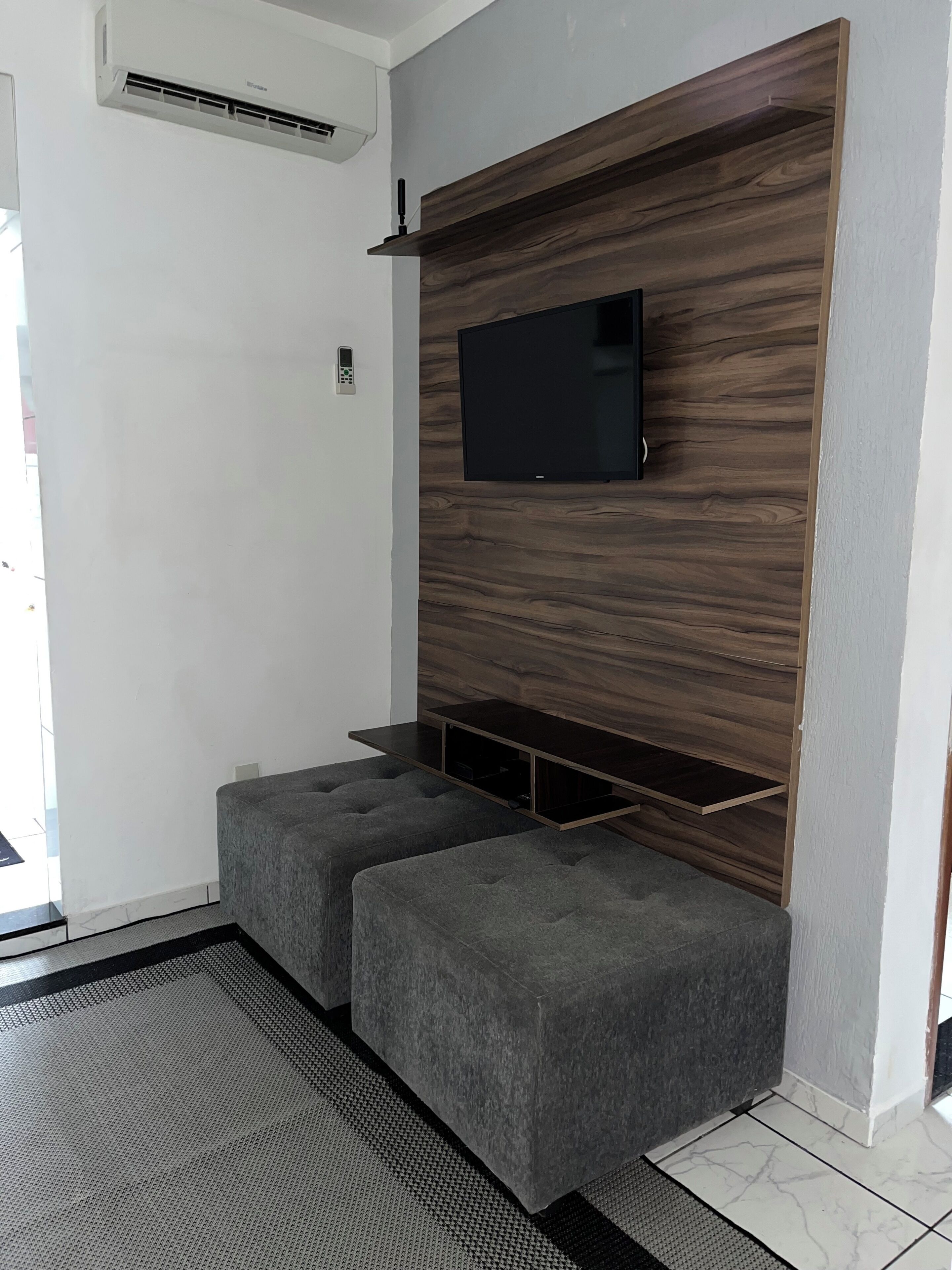Ein modernes Wohnzimmer mit einem Flachbildfernseher, der an einer Holzpaneelenwand angebracht ist, zwei grauen Sitzgelegenheiten und einem kleinen Regal unter dem Fernseher.