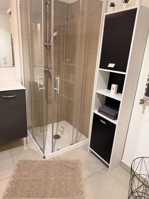 A shower with a glass door, a white shelf unit with a folded towel, and a beige rug. - Detached studio apartment across from Riquier Station, with a beautiful view of the hills of Nice (Nice)