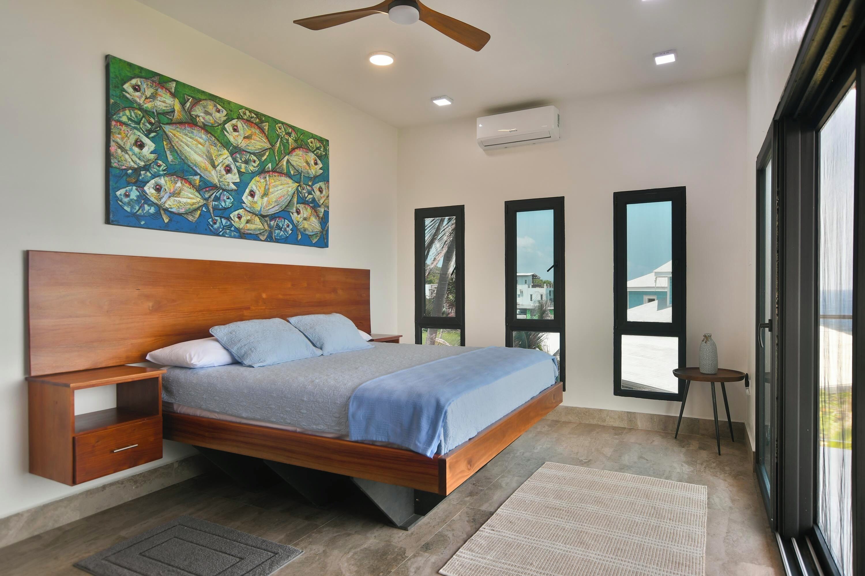 A modern bedroom with a wooden bed, a nightstand, a large painting, and a view through the windows.