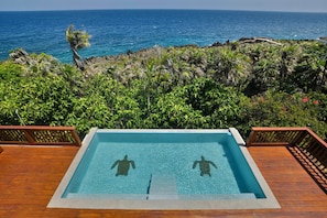 A rectangular infinity pool with clear blue water, surrounded by wooden deck and lush greenery, overlooking a rocky coastline and the ocean. - Casa Tranquilidad 3-bed by Island House Vacation Rentals (West Bay)