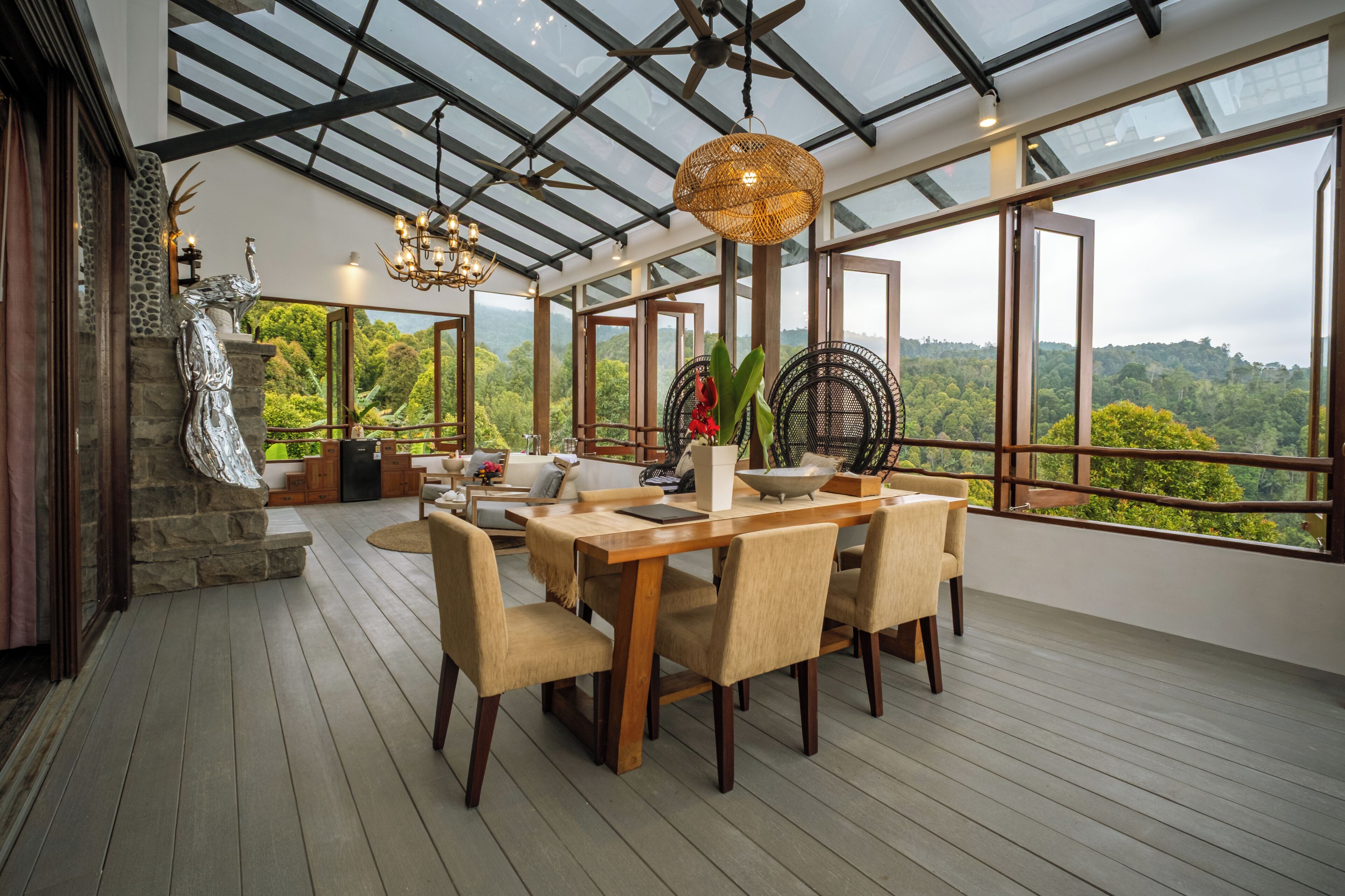 A spacious dining area with a wooden table and chairs, a hanging light fixture, and a view of lush greenery through large windows.