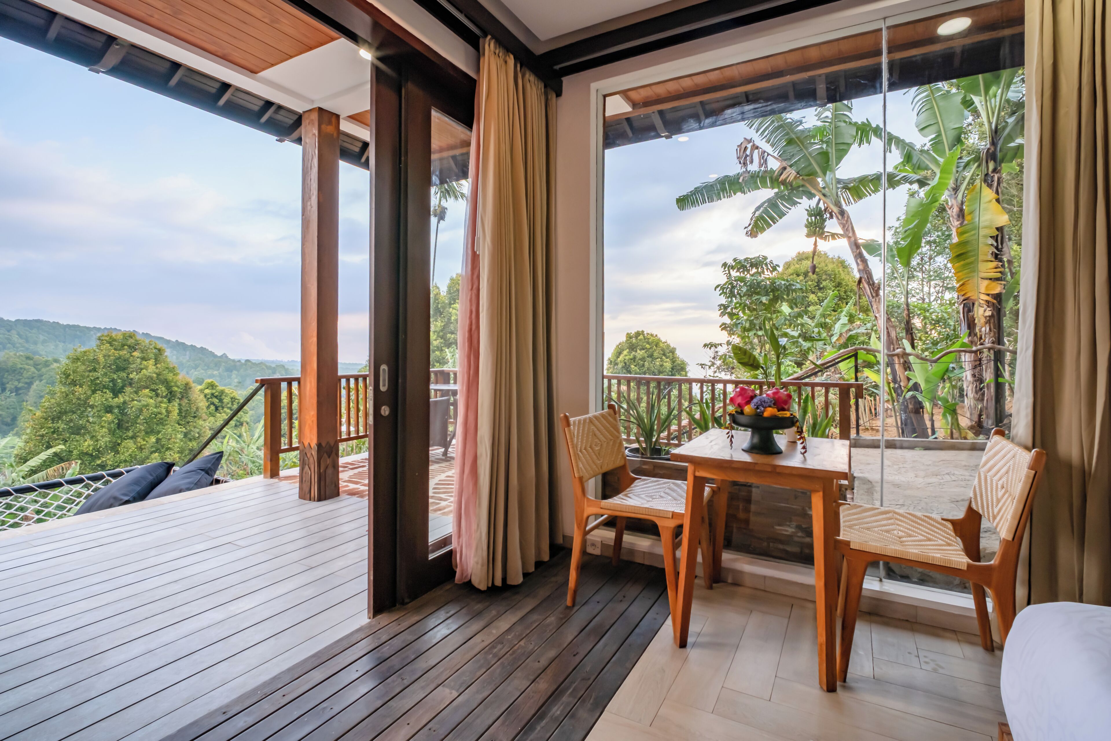 A balcony with wooden flooring, a table with chairs, and a view of lush greenery and hills.