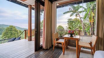 A balcony with wooden flooring, a table with chairs, and a view of lush greenery and hills.