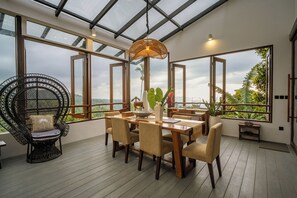 A dining area with a wooden table and chairs, a wicker chair, and a view of the outdoors through large windows.