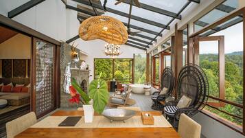 Une pièce spacieuse avec une grande table en bois, des sièges confortables et une vue sur une végétation luxuriante grâce à de grandes fenêtres.