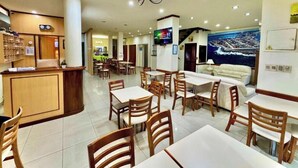 Un hall de hotel con mostrador de recepción, zona de comedor con mesas y sillas, un sofá y un mural grande en la pared.