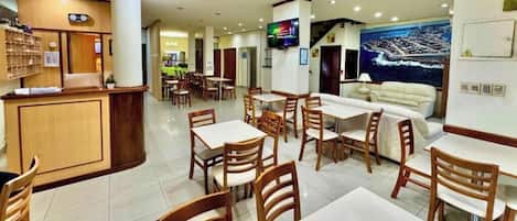 A hotel lobby with a reception desk, dining area with tables and chairs, a sofa, and a large mural on the wall.