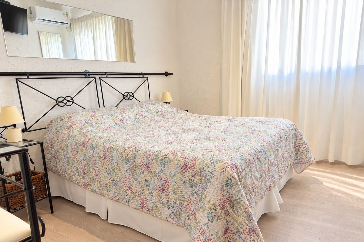 Un dormitorio con cama, lámparas de noche, un espejo y una ventana con cortinas.