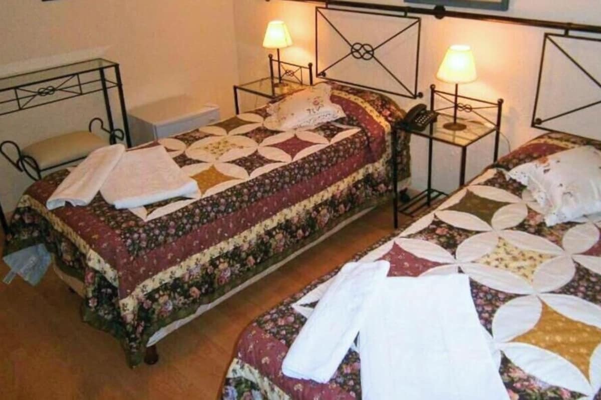 A room with two single beds, each with a metal frame and a floral-patterned bedspread. There are bedside tables with lamps and a wall-mounted shelf above the beds.