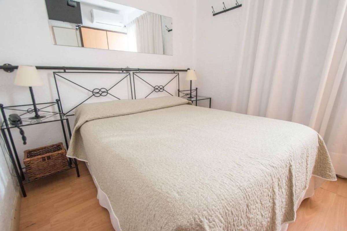 A single bed with a headboard, bedside lamps, a small wicker basket, and a mirror on the wall.