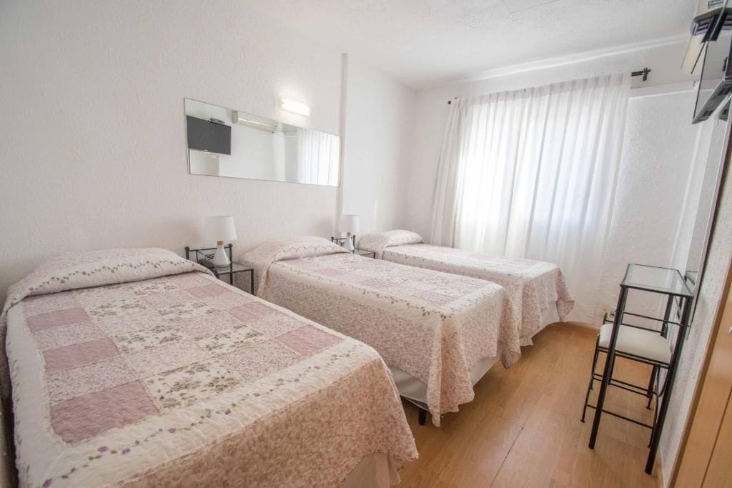 Habitación de hotel con dos camas, un espejo, una mesita de noche con lámpara y una ventana con cortinas.
