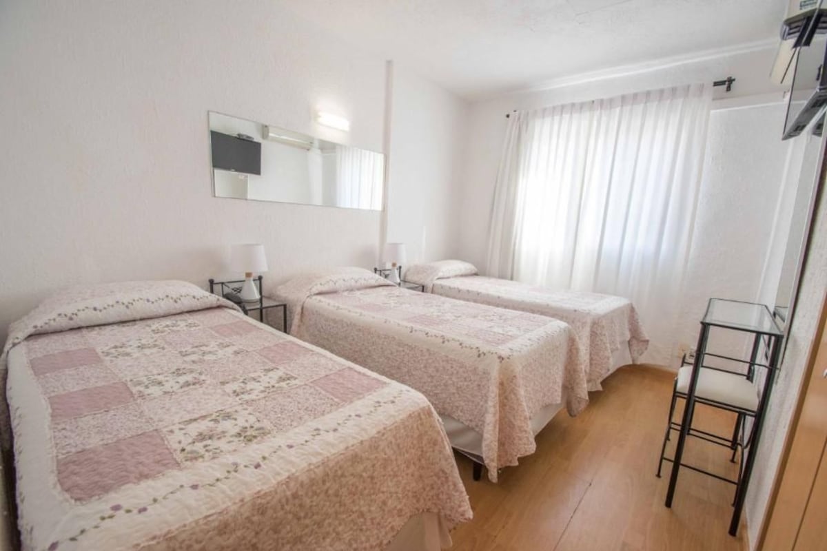 A hotel room with two beds, a mirror, a bedside table with a lamp, and a window with curtains.