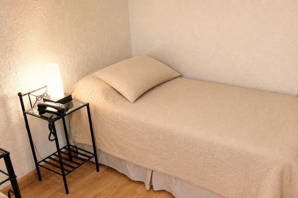 A single bed with a beige bedspread and pillows, a bedside table with a lamp, and a telephone.