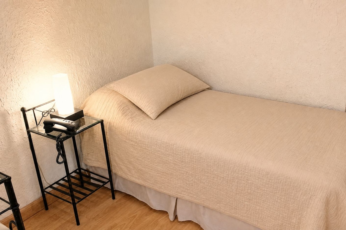 A single bed with a beige bedspread and pillows, a bedside table with a lamp, and a telephone.