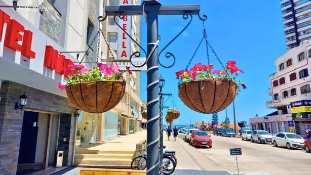 Vista de la calle con un letrero de hotel, cestas de flores colgantes y coches estacionados.