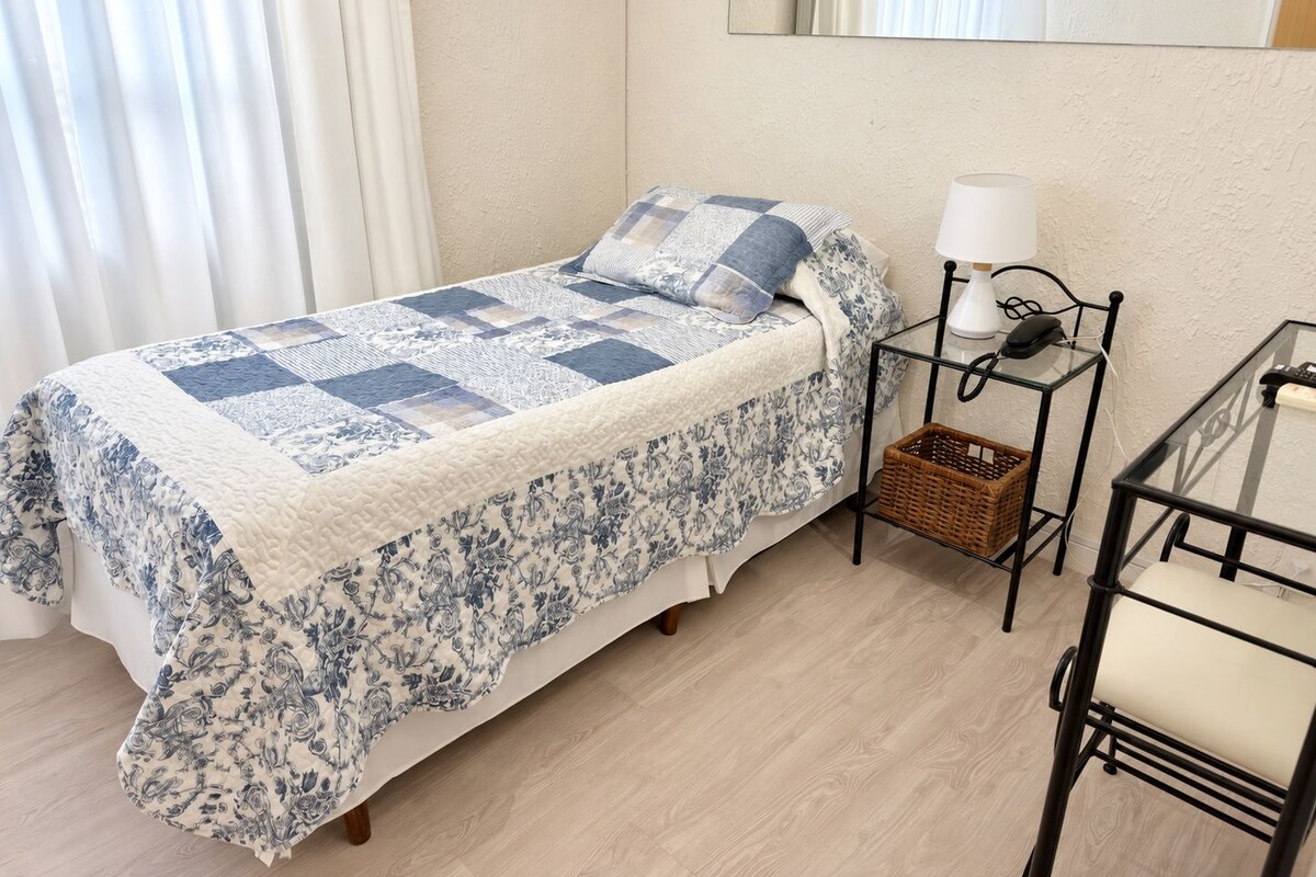 Una cama individual con colcha, una mesita de noche con lámpara, una cesta de mimbre y una silla.
