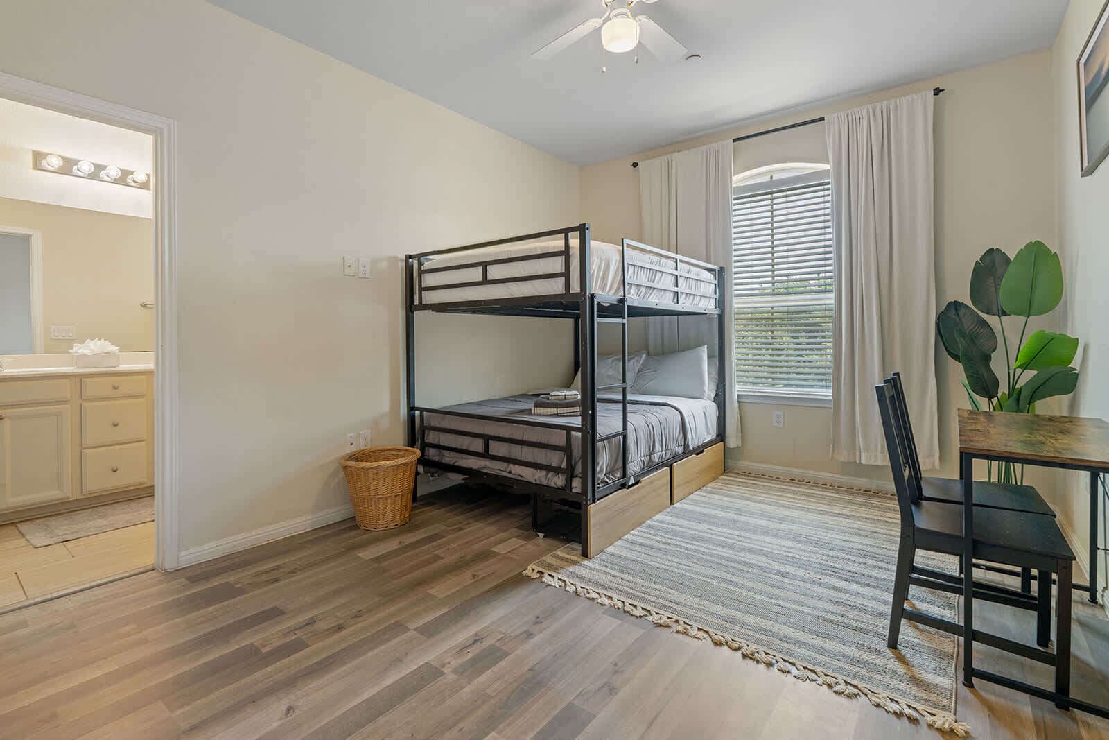 Une chambre avec un lit superposé, une petite table avec une chaise, un panier et une fenêtre avec des rideaux.