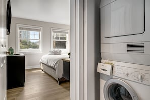 Una lavandería con una lavadora Bosch, una cama con edredón blanco y una ventana con persianas.
