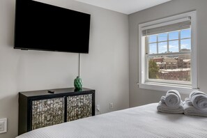 Una camera da letto con un letto, una televisione a parete, un comò con un vaso decorativo, una finestra vista edifici e asciugamani arrotolati sul letto.