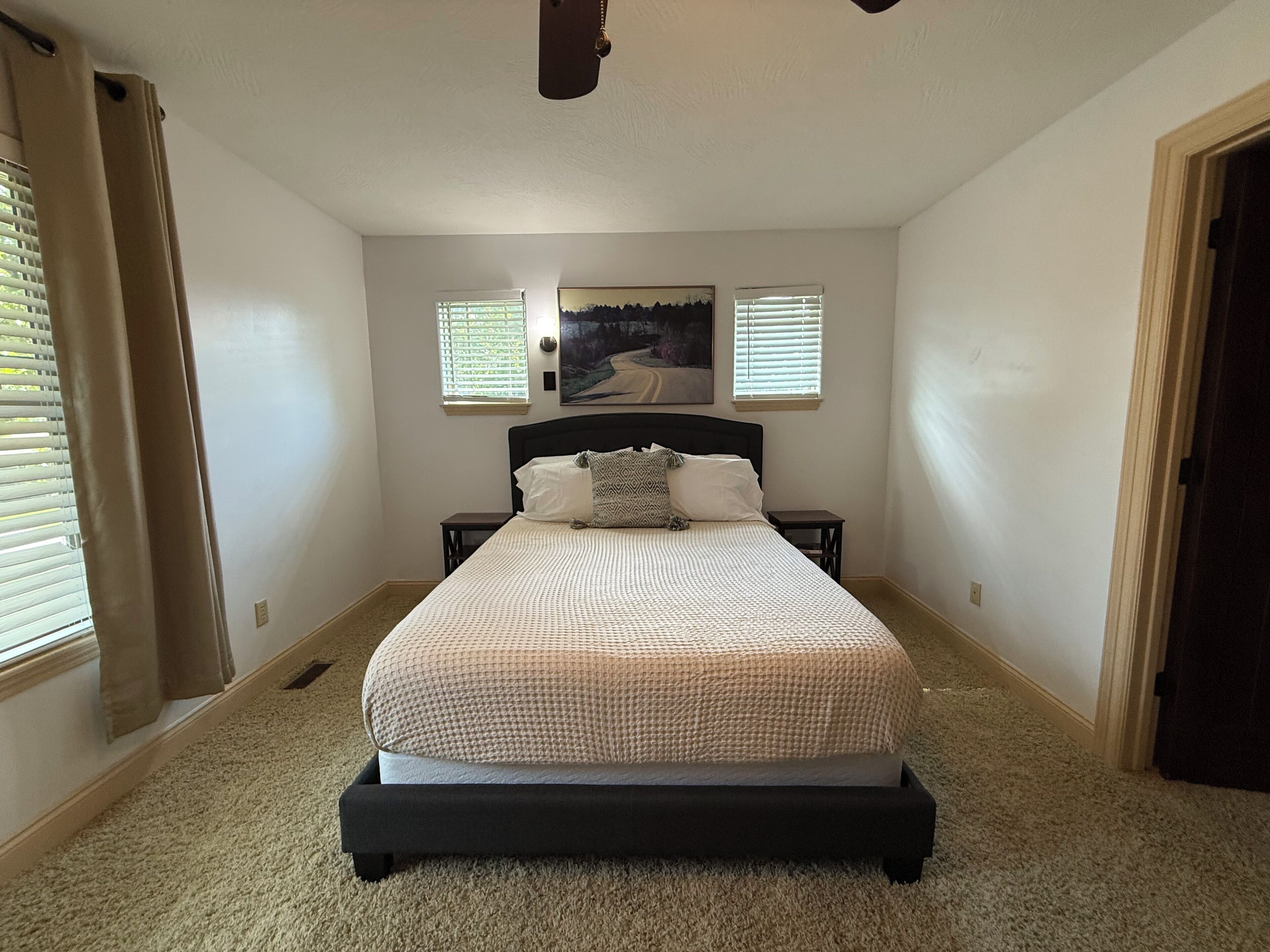 A bedroom with a bed, a nightstand, a painting, and two windows with blinds.