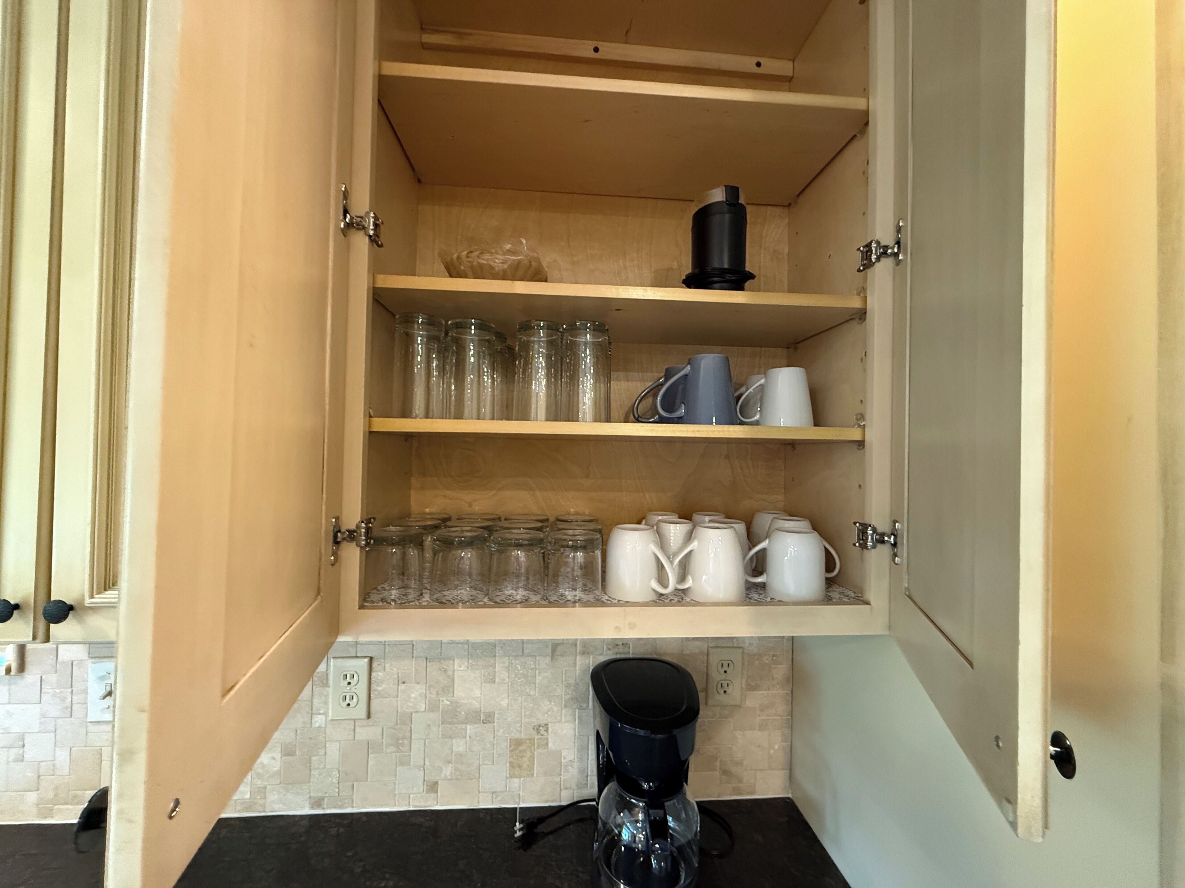 A kitchen with a coffee maker on the counter, a cabinet with shelves holding mugs and glass jars.