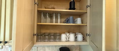 A kitchen with a coffee maker on the counter, a cabinet with shelves holding mugs and glass jars.