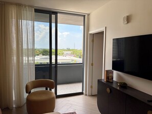 Una habitación con puerta corrediza de vidrio que da a un balcón, un televisor de pantalla plana en la pared, una silla marrón y un mueble de madera oscura.
