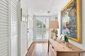 A hallway with white walls, a wooden bench, a lamp, a framed painting, and a door with white shutters. - Heron Marsh 108 Beautiful view, Litchfield by the Sea (Pawleys Island)