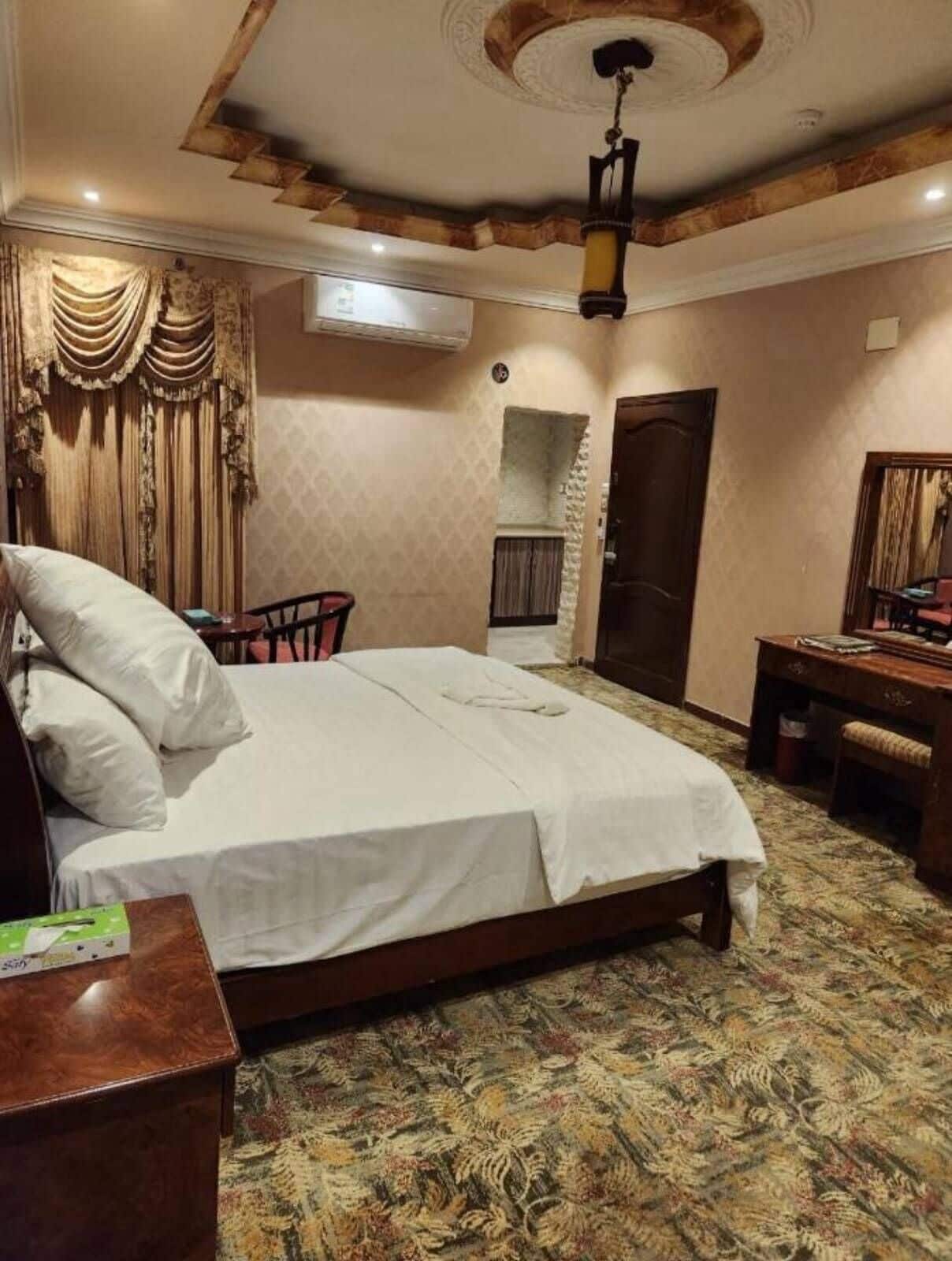 A hotel room with a bed, a desk, a chair, an air conditioning unit, and a patterned carpet.