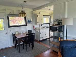 A compact kitchen with white cabinets, a dining table with chairs, a hanging light fixture, and a microwave on a shelf. - Waterfront peaceful 2-bedroom cottage in charming Freeport perfect for relaxing (Freeport)