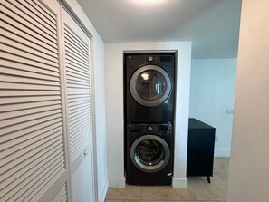 Uma lavanderia com máquina de lavar e secadora de abertura frontal, um armário branco fechado e um banco de cor escura.
