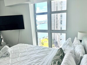 A hotel room with a bed, a TV mounted on the wall, and a view of a cityscape through large windows. - Luxury Brickell Condo - Bay View - Garage Parking (Miami)