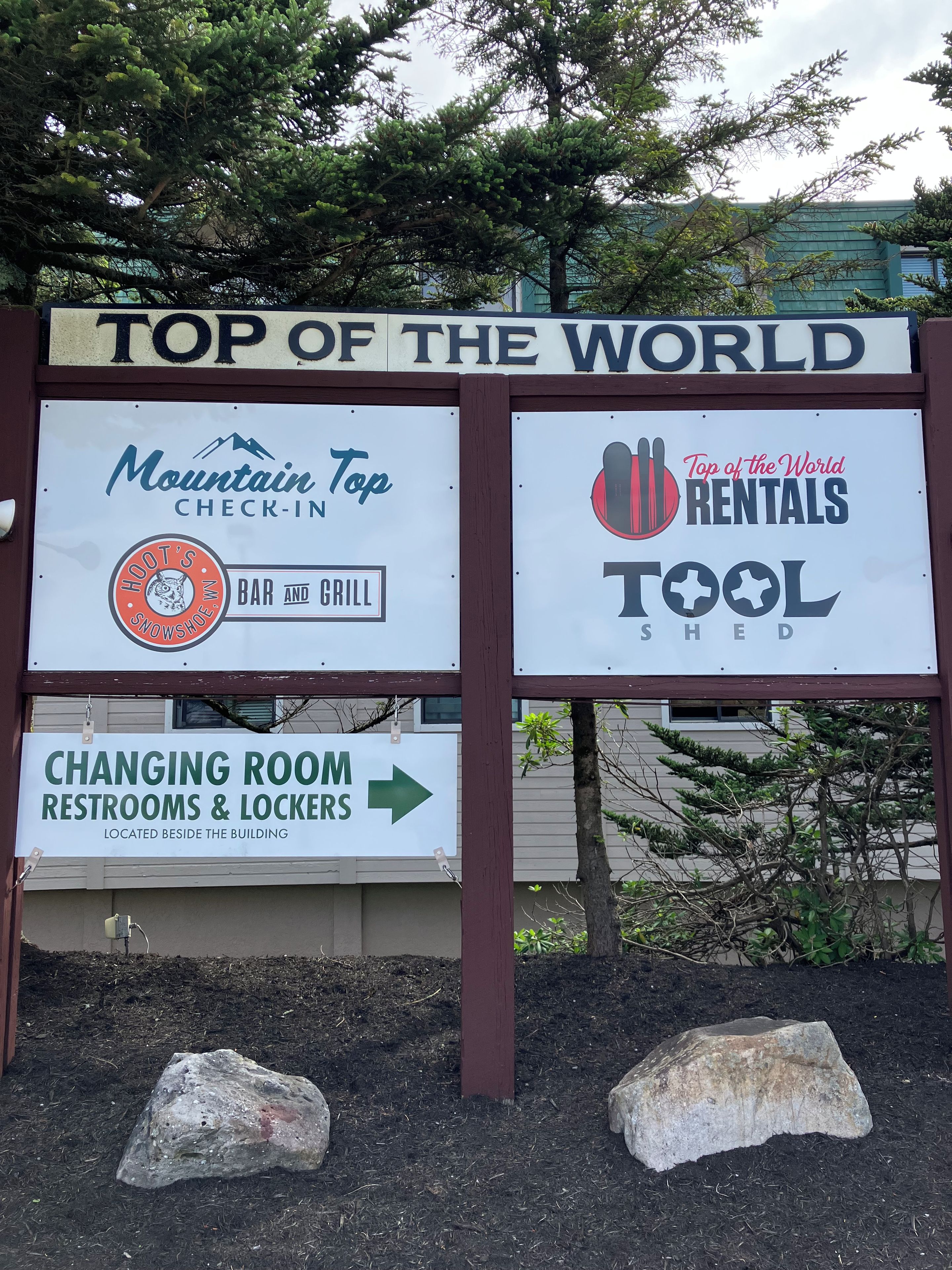 A signboard with multiple advertisements including 'Mountain Top Check-In,' 'Hoot's Snowshoe Bar and Grill,' 'Top of the World Rentals,' and directions to 'Changing Room Restrooms & Lockers.'