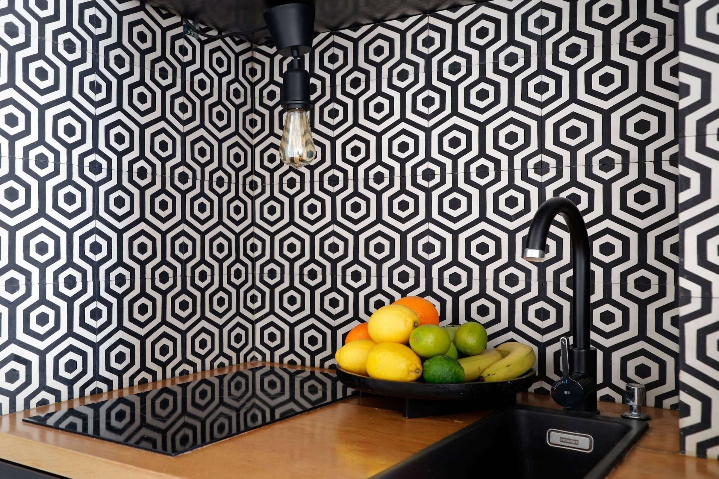 A kitchen with a black and white hexagonal tile backsplash, a black faucet, and a bowl of fruit on the counter.