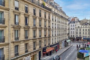 Ein mehrstöckiges Gebäude mit einer Reihe von Balkonen, ein Laden mit einer Pizzeria und eine Straße mit Fahrzeugen und Fußgängern.