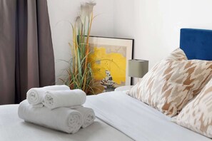 Ein ordentlich bezogenes Bett mit aufgerollten weißen Handtüchern, einem blauen Kopfteil und gemusterten Kissen.