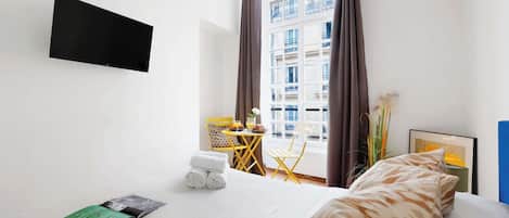 Hotellihuone, jossa on sänky, seinälle asennettu taulutelevisio, pieni pöytä ja tuoli sekä ikkuna, jossa on verhot.