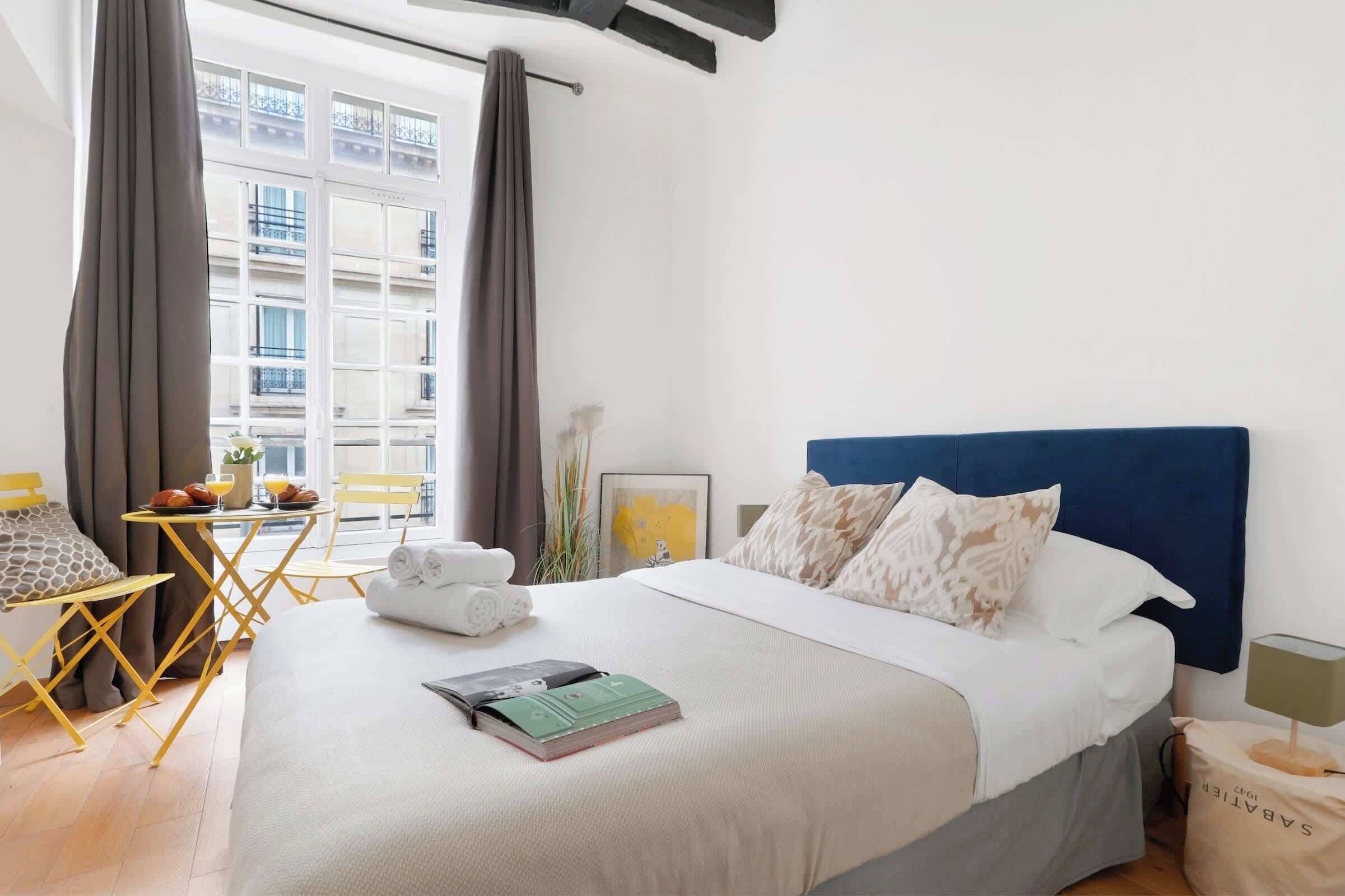 A hotel room with a bed, a small table with breakfast items, a chair, and a view of buildings through large windows.