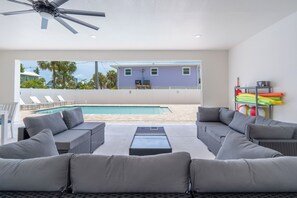 A modern outdoor living area with a pool, grey sofas, and a ceiling fan. - 🌴Seaside Cottage | Steps to the Beach 🏖️ Gulf Sunsets & Island Bliss 🌅 (Fort Myers Beach)
