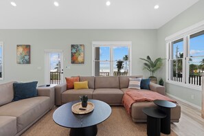 A spacious living room with a beige sectional sofa, a round coffee table, and a large window offering a view of the beach. - 🌴Seaside Cottage | Steps to the Beach 🏖️ Gulf Sunsets & Island Bliss 🌅 (Fort Myers Beach)