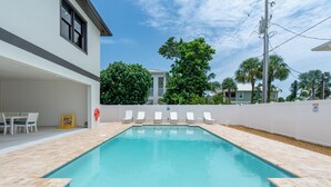 Ein rechteckiger Swimmingpool mit Fliesenränder, umgeben von einem weißen Zaun, und einer überdachten Terrasse mit weißen Stühlen.