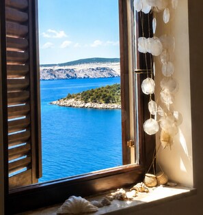 Ein Fenster mit Blick auf das blaue Meer und eine felsige Insel, Holzjalousien und eine Wanddekoration mit hängenden Objekten.