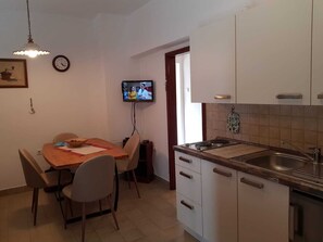 Eine kompakte Küche mit weißen Schränken, einem Esstisch mit Stühlen, einem an der Wand montierten Fernseher und einem Fenster für natürliches Licht.