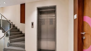 An elevator with a digital display, a staircase with glass railings, and a wooden door with a circular design. - Classic Simran Raghuveer Para (Rajkot)