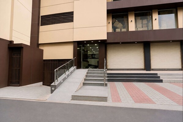 A building with a staircase leading to a door, a closed shutter, and a paved area with a metal railing.