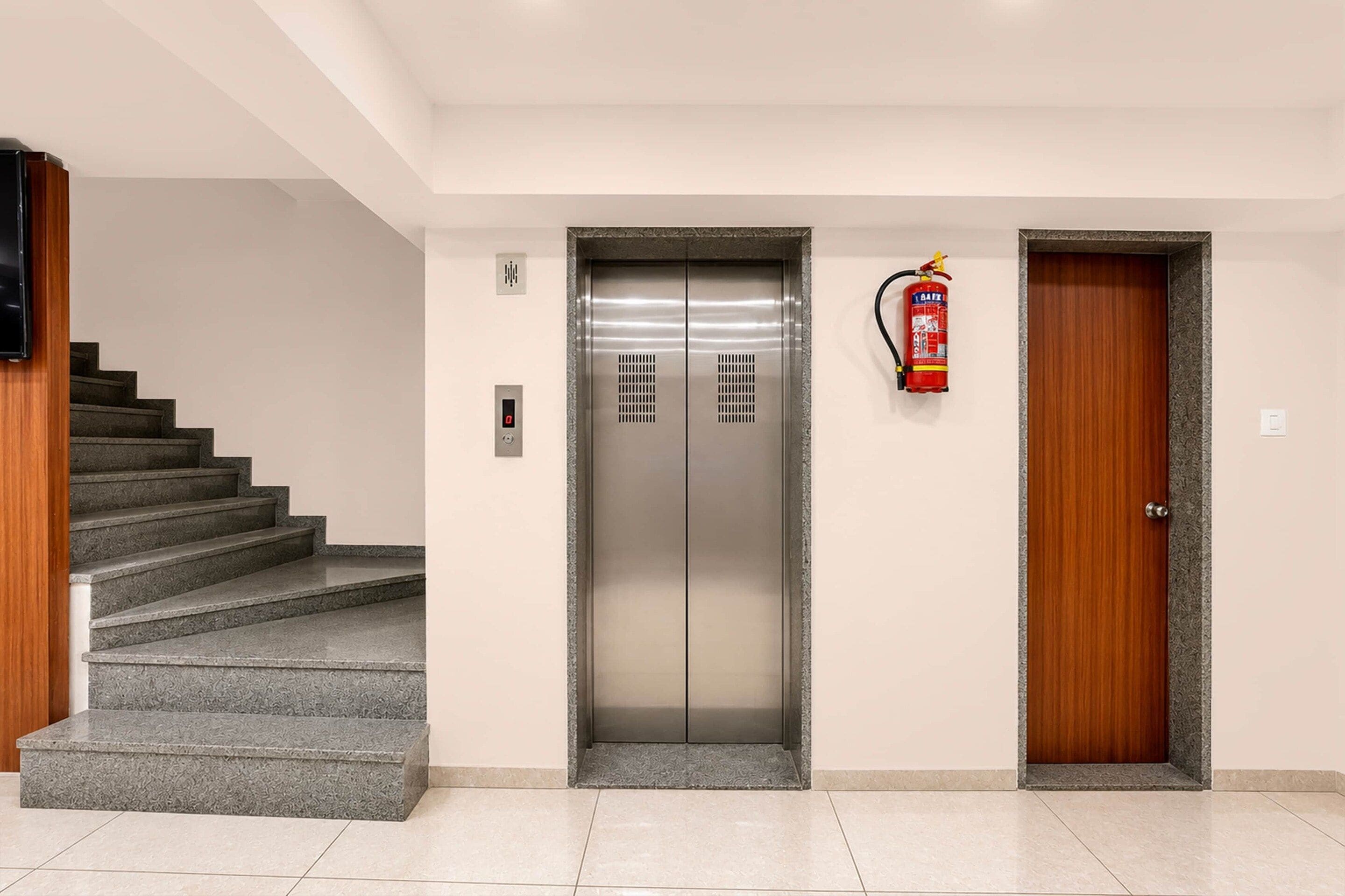 A modern elevator with a digital display, a fire extinguisher mounted on the wall, and a wooden door to the right.