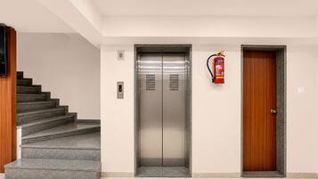 A modern elevator with a digital display, a fire extinguisher mounted on the wall, and a wooden door to the right.